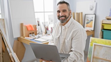 Gülümseyen, sakallı, gri saçlı, resim stüdyosunda dizüstü bilgisayar kullanan olgun bir İspanyol.