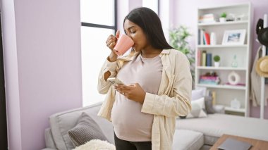 Genç, kaygısız İspanyol kadın, evinde hamileliğin mutluluğunu yaşıyor, sıcak kahveyi yudumlarken rahat oturma odasında akıllı telefonundan sabah mesajları okuyor.