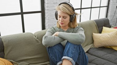 Gündelik giysiler içinde, kulaklıklı müzikten hoşlanan, evdeki bir kanepede oturan, sakin bir ifadeyle oturan genç sarışın kadın..