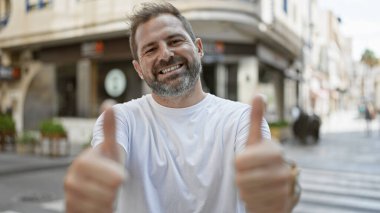 Şehrin bulanık arka planına sahip bir sokak ortamında gülümseyen sakallı adam başparmağını kaldırıyor..