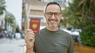 Beyaz saçlı, kendinden emin orta yaşlı bir adam, bir şehir parkında duruyor, keyifle Fransız pasaportunu tutuyor, sakin bir Avrupa tatiline hazır..