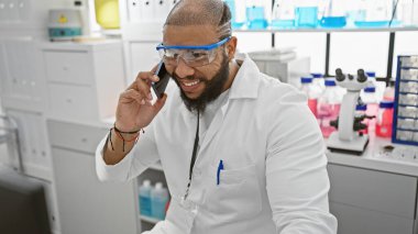 Sakallı yakışıklı Afro-Amerikan adam laboratuvar ortamında telefonla konuşurken gülümsüyor..