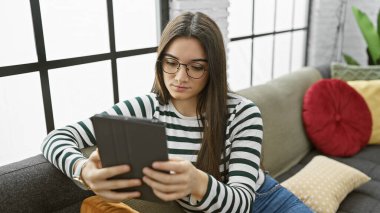 Gözlüklü, rahat bir kanepede tablet okuyan odaklanmış genç bir kadın..