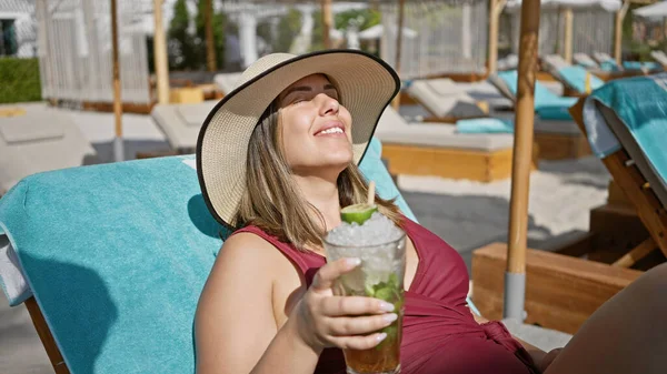 Güneş şapkalı gülümseyen bir kadın lüks bir tatil köyünde plaj sandalyesinde uzanırken mojito içiyor..