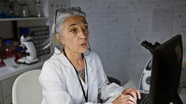 Middle age grey-haired lady scientist, focused face illuminated by the computers glow, diligently working on breakthrough research in a quiet, dimly lit lab