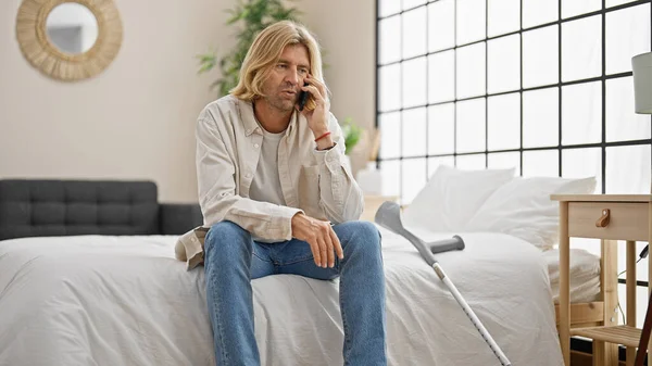 Blond Man Talks Phone Sitting Bed Crutches Bright Bedroom — Stock Photo, Image