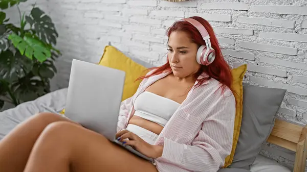 红头发美女舒适地蜷缩在床上 一边穿着舒适的睡衣 笔记本电脑和耳机 一边认真地在网上播放着曲调 — 图库照片