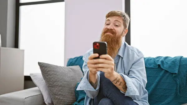 Handsome young redhead man cheerfully using his smartphone while sitting on the sofa in the living room of his home, spreading joy and positive vibes through texting and the online world