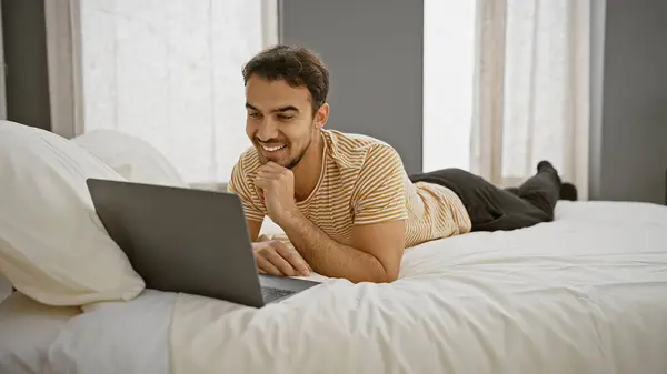 A smiling young man lies on a bed with a laptop in a bright bedroom, indicating comfort and leisure at home.