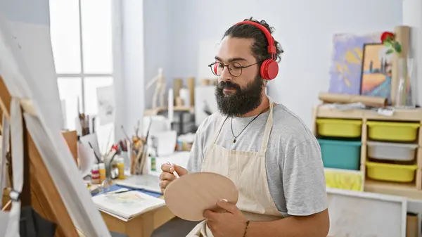 Handsome bearded hispanic man with headphones painting in a bright art studio