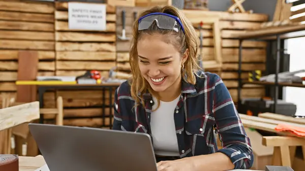 Gorgeous young hispanic woman carpenter utilizing laptop for online woodworking business in her cozy carpentry workshop, safeguarded by security glasses