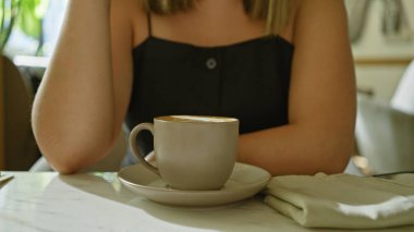 Genç bir esmer kadın kafeterya masasında cappuccino içiyor, keyfi ve modern yaşam tarzını somutlaştırıyor..