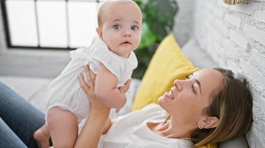 Mutlu anne yatakta uzanıyor, mutlu küçük kızını havada tutuyor, gülümsüyor ve sıcak, sade yatak odasında oynarken eğleniyor..