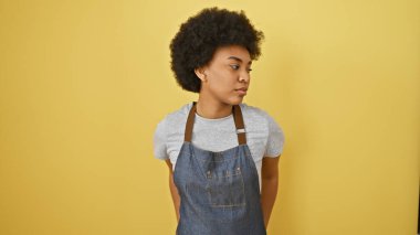 Sarı arka planda kıvırcık saçlı, kot önlük giyen düşünceli Afrikalı Amerikalı bir kadının portresi..