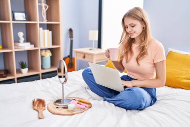 Young blonde woman using laptop drinking coffee sitting on bed at bedroom