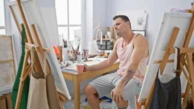 Sakallı genç bir adam bir sanat stüdyosunda oturmuş yaratıcı bir kaosun ortasında kahve fincanı tutuyor..