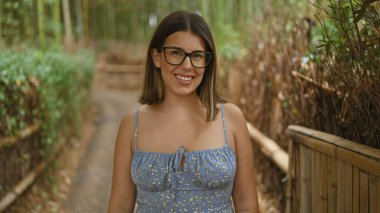 Neşeli, güzel İspanyol kadın, kendinden emin bir şekilde poz veriyor, gözlük takıyor, Kyoto 'nun bambu ormanının yeşilliğinin ortasında duruyor.