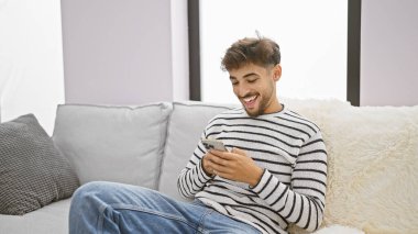 Neşeli genç bir Arap adam, koltuğunda rahat bir şekilde otururken akıllı telefonuyla sevinçle mesaj atıyor. Evinin sıcak, rahat oturma odasında mutluluk saçıyor.