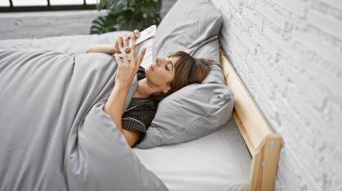 Yatakta uzanmış akıllı telefon kullanan genç, güzel İspanyol kadın.