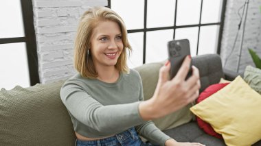 Sarı saçlı yetişkin bir kadın rahat bir oturma odasındaki kanepede otururken telefonuna selfie çekiyor..