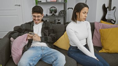 Modern oturma odasındaki kanepede ciddi bir kadının yanında akıllı telefon kullanan gülümseyen adam.