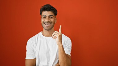 Sakallı, yakışıklı, genç, İspanyol bir adam kıpır kıpır kırmızı bir duvara yaslanmış gülümseyip el kol hareketleri yapıyor..