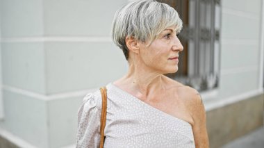 Şık, olgun, kır saçlı, şehir ortamında düşünceli bakışları kaçıran bir kadın..