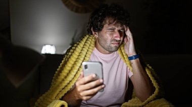 Baş ağrısı olan sıkıntılı İspanyol bir adam gece evinde akıllı telefon kullanıyor, sarı bir battaniyeye sarılmış..