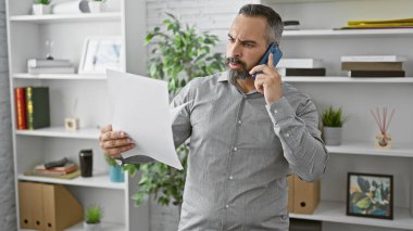 Modern bir ofis ortamında telefon görüşmesinde profesyonellik ve konsantrasyon duygusunu aktarırken gri sakallı, olgun İspanyol bir adam belgeleri tartışıyor..