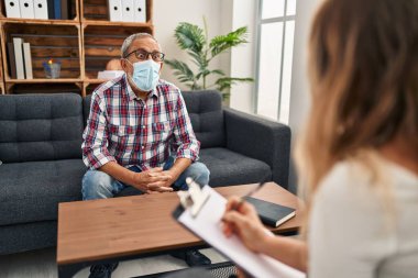 Korkmuş kıdemli bir adam, yüzü inançsızlıkla dolu, psikoloji kliniğinde şaşkın bir şekilde oturuyor, tıbbi maske destansı bir sürprizle ağzı açık.