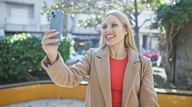 Gülümseyen genç sarışın kadın yeşillik ve sokak geçmişiyle dışarıda selfie çekiyor..