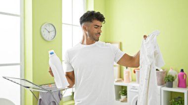 Yakışıklı İspanyol bir adam aydınlık bir odada çamaşır yıkıyor, elinde deterjan tutuyor ve beyaz bir gömleği inceliyor..