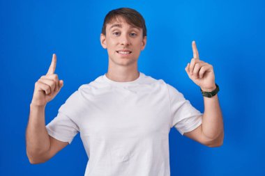 Mavi arka planda duran beyaz tenli sarışın adam şaşırmış, şaşırmış ve parmaklarıyla işaret ediyor ve kollarını kaldırıyordu.. 