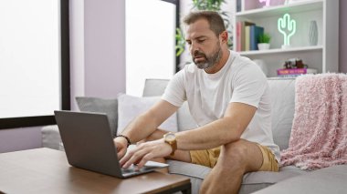 Modern oturma odasında dizüstü bilgisayar kullanan, ev işleri yapan, beyaz saçlı, olgun İspanyol bir adam..