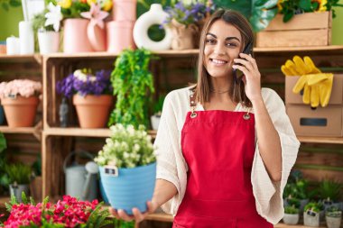 Young beautiful hispanic woman florist talking on smartphone holding plant at flower shop