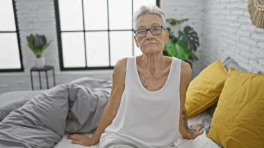 Ciddi görünümlü, gri saçlı yaşlı kadın rahat bir şekilde rahat yatağında dinleniyor, derin düşünceler içinde, sade yatak odasının sıcaklığında, soluk bir sabah ışığı içeri sızıyor.