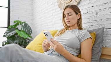 Genç, çekici sarışın kadın, rahat bir şekilde yatağında oturuyor, rahat bir sabah rahat bir yatak odasında akıllı telefonunu kullanmaya dalmış.