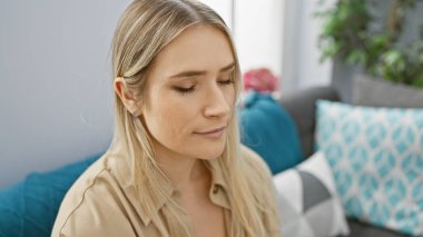 Güzel sarışın kadın şüphesiz meditasyon yaparken derin derin düşünür, evdeki kanepede oturur, endişeli ifadeler kapalı gözlerine yansıyor, oturma odasının arka planında iç mekan portresi