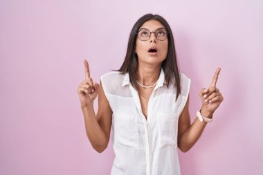 Pembe arka planda duran esmer genç bir kadın şaşırmış ve şaşırmış bir şekilde yukarı bakıyor ve parmaklarıyla işaret ediyor ve kollarını kaldırıyor.. 