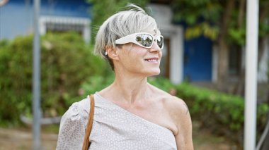 Gri saçlı ve güneş gözlüklü olgun bir kadın yeşil bir şehir parkında gülümsüyor..