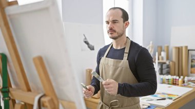 Önlüklü, odaklanmış, sakallı bir adam bir sanat stüdyosunda duruyor, elinde bir sehpanın yanında fırça tutuyor..