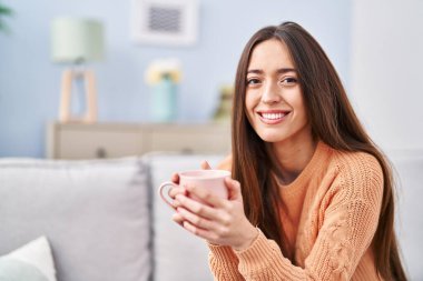 Genç ve güzel İspanyol kadın evdeki kanepede oturup kahve içiyor.