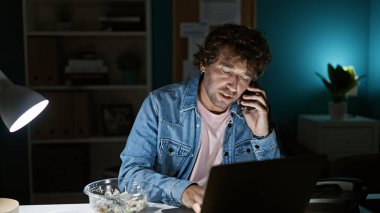 İspanyol adam geceleri ofiste birden fazla iş yapıyor, dizüstü bilgisayar kullanıyor ve telefonla konuşuyor..