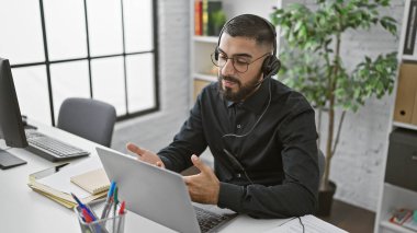 Modern ofiste kulaklık takan sakallı adam iş sırasında bilgisayarda iletişim kuruyor.