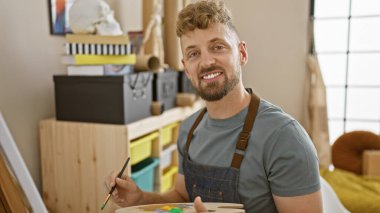 Mavi gözlü, sakallı, sanat stüdyosunda önlük takan, fırçayla resim yapan neşeli beyaz bir adam.