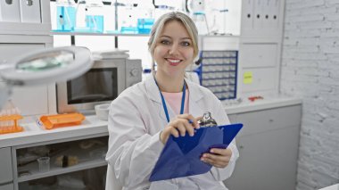 Laboratuvar önlüklü genç sarışın bir kadın elinde bir pano, parlak bir laboratuvar ortamında gülümsüyor..