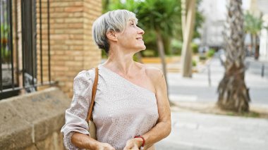 Gri kısa saçlı olgun bir kadın hareketli bir caddede dikilirken yukarı bakar..
