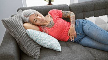 Endişeli orta yaşlı kadın, zarif gri saçlı, dinlenirken şiddetli karın ağrısı çeken, mutsuzca oturma odasındaki kanepede uzanan.