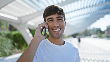 Güneşli parkta, kendine güvenen genç bir İspanyol adam duruyor, gülüşü ışıldıyor. Akıllı telefonundaki bir çağrıya kendini kaptırdı. Havalı bir resim. Açık havada saf mutluluğun portresi.