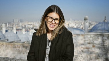 Bulanık İstanbul şehir manzarasının önünde gözlüklü, gülümseyen genç kadın, kentsel Türk kültürünü vurguluyor